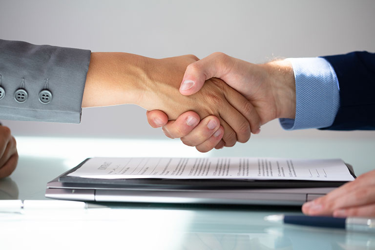 People shaking hands with a laptop and paperwork on the table.